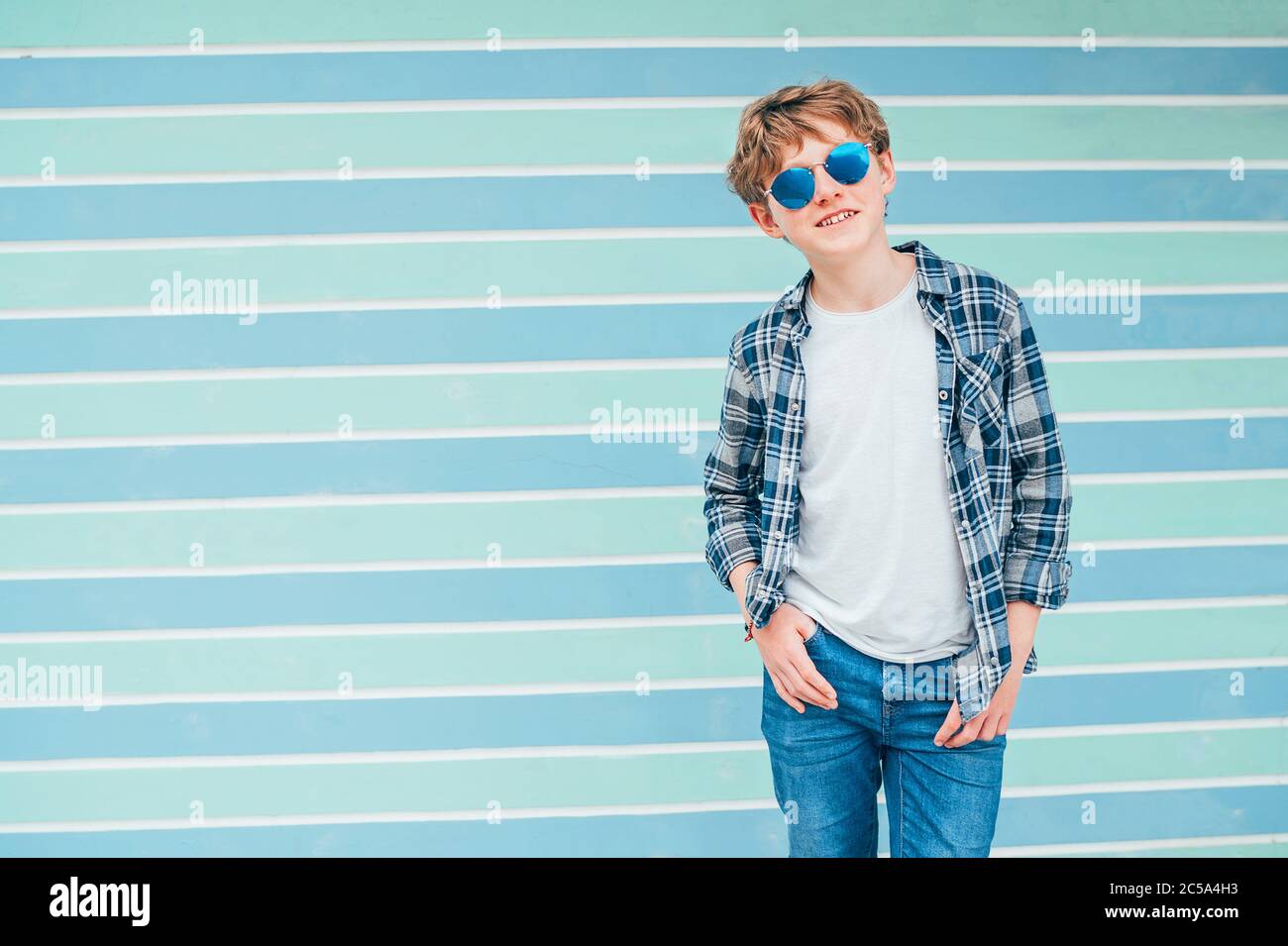 Kaukasische blonde Haare 12 Jahre alter Teenager Junge Mode-Portrait gekleidet weißes T-Shirt mit karierten Hemd in blauer Sonnenbrille mit türkisblauem Rücken Stockfoto