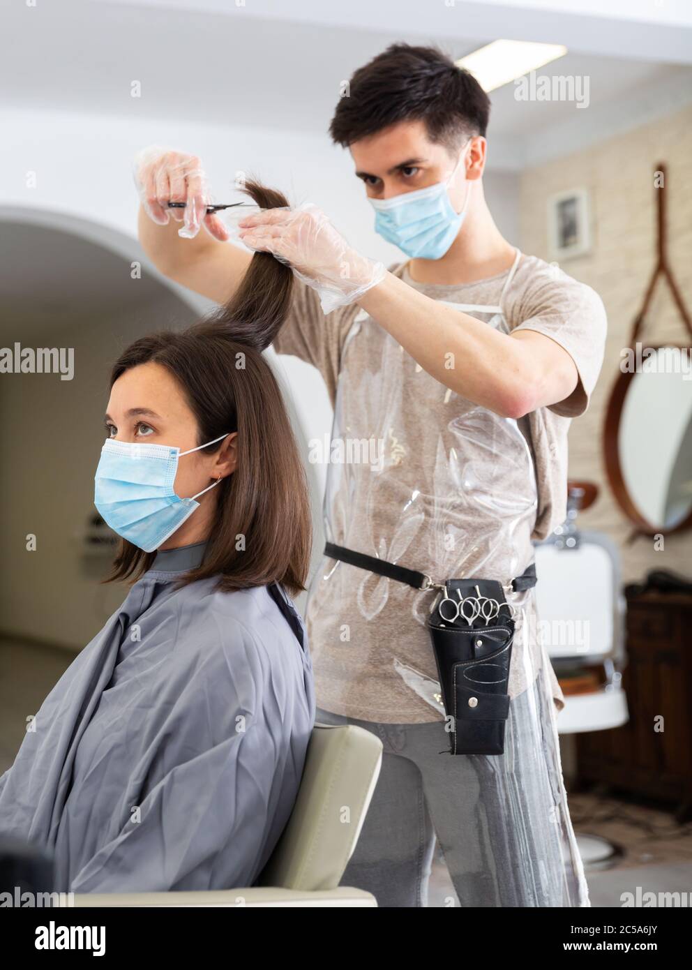 Positive männliche Friseur in Schutzmaske und Handschuhe machen Haar Styling für junge Frau, Arbeitstag im Schönheitssalon während Pandemie-Situation Stockfoto