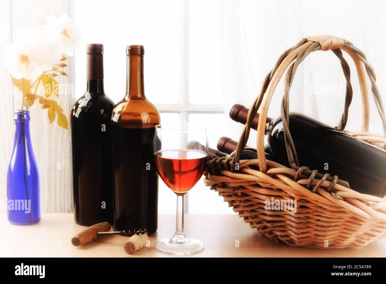 Wein Stillleben mit einem Korb von Flaschen und ein Glas Rotwein und weiße Rosen in einer Vase auf einem Tisch vor einem Fenster mit Vorhängen. Horizontal f Stockfoto