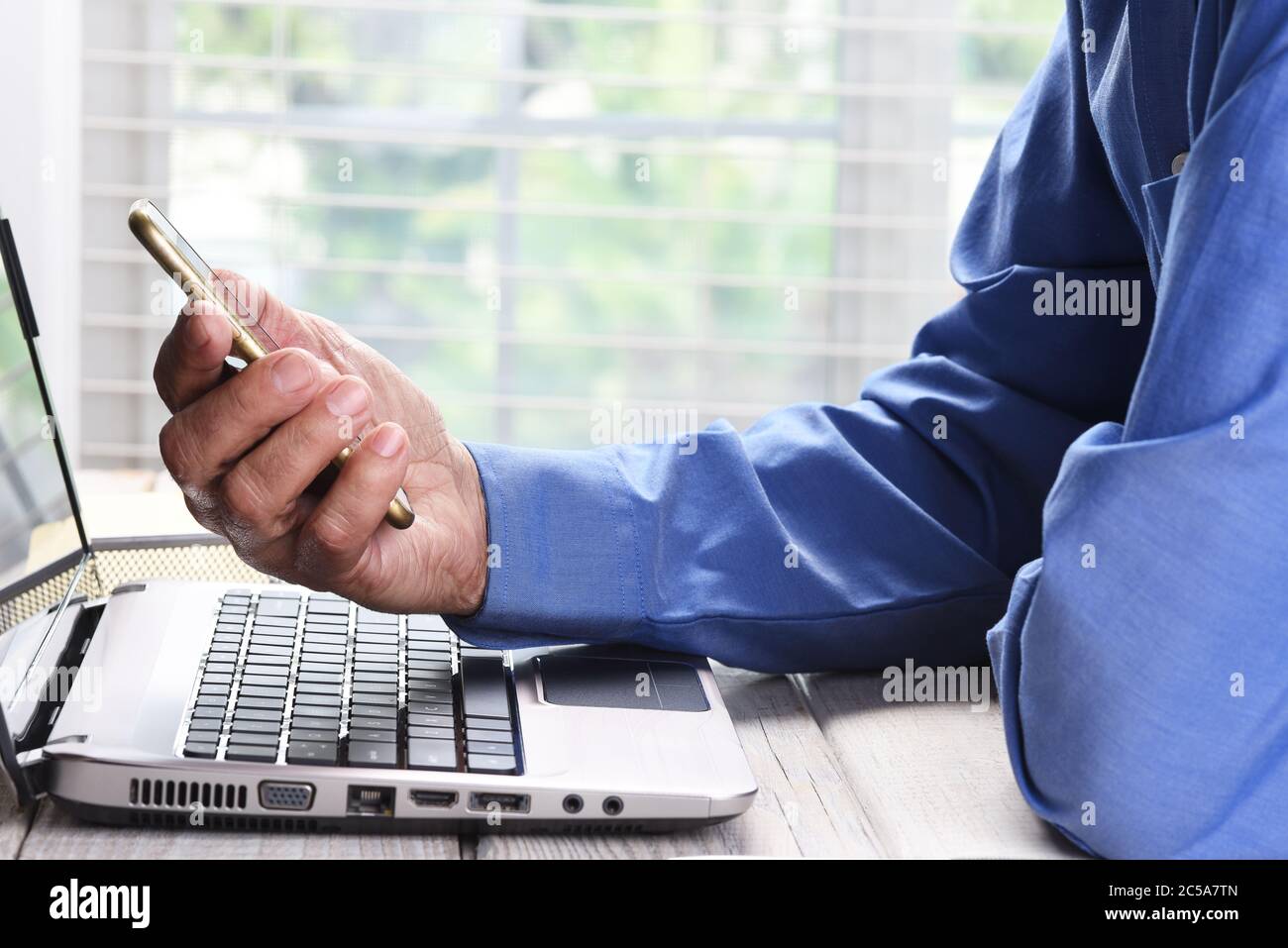 Nahaufnahme eines Mannes, der ein Mobiltelefon in einer Hand über einen Laptop auf dem Schreibtisch des Heimbüros hält. Stockfoto