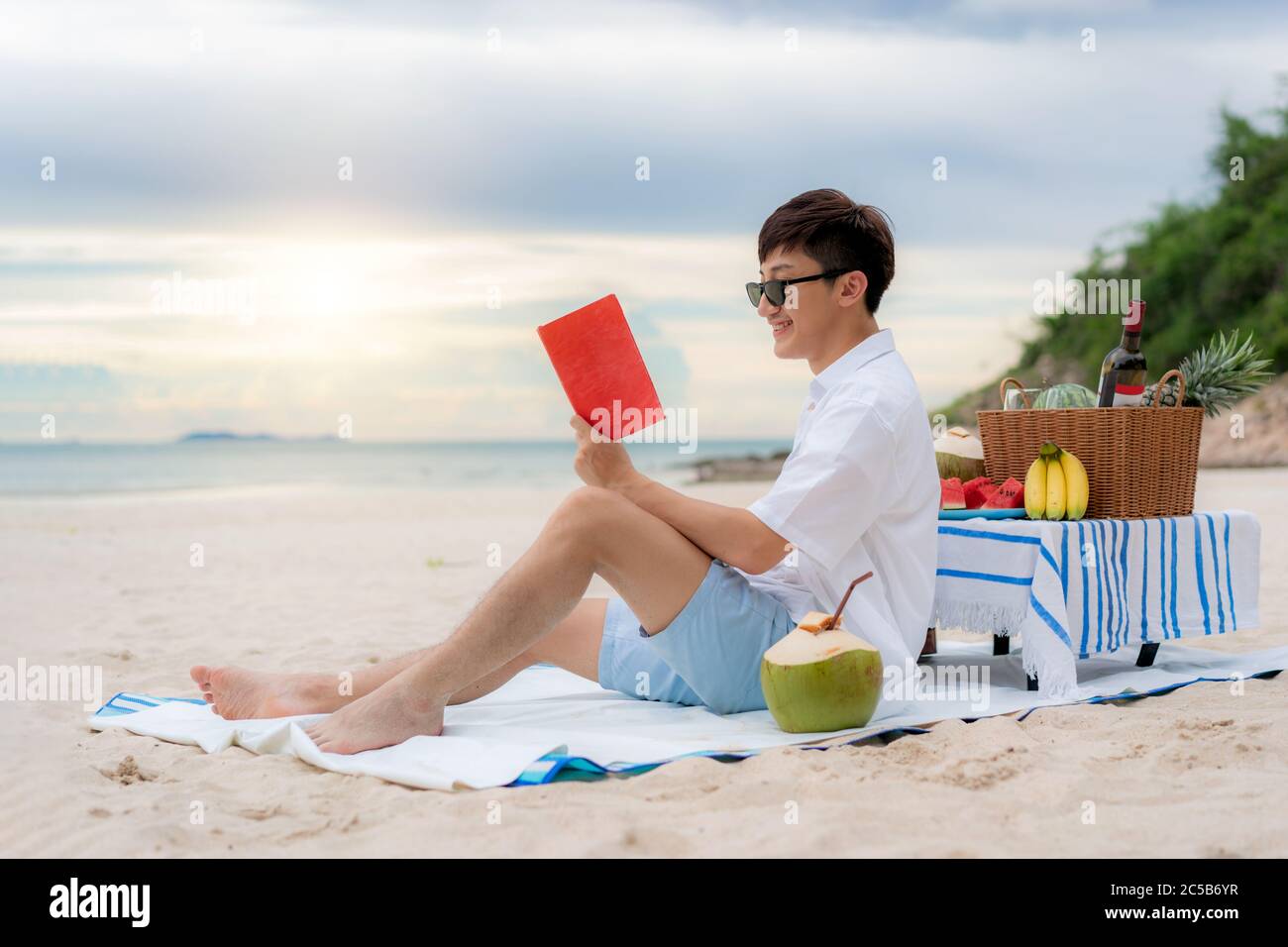 Junge asiatische Mann tragen Sonnenbrille ist mit einem Buch im weißen Sandstrand und in der Nähe des Meeres mit tropischen Früchten im Hintergrund entspannen. Sommer, Urlaub Stockfoto