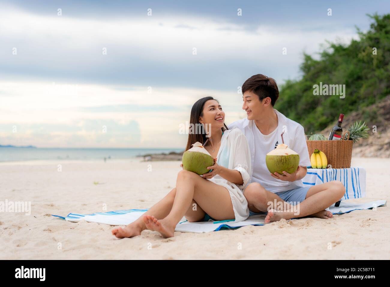 Asiatisches junges Paar auf der Picknickdecke sitzen und Kokoswasser am Strand und in der Nähe des Meeres mit tropischen Früchten im Hintergrund trinken. Sommer, holid Stockfoto