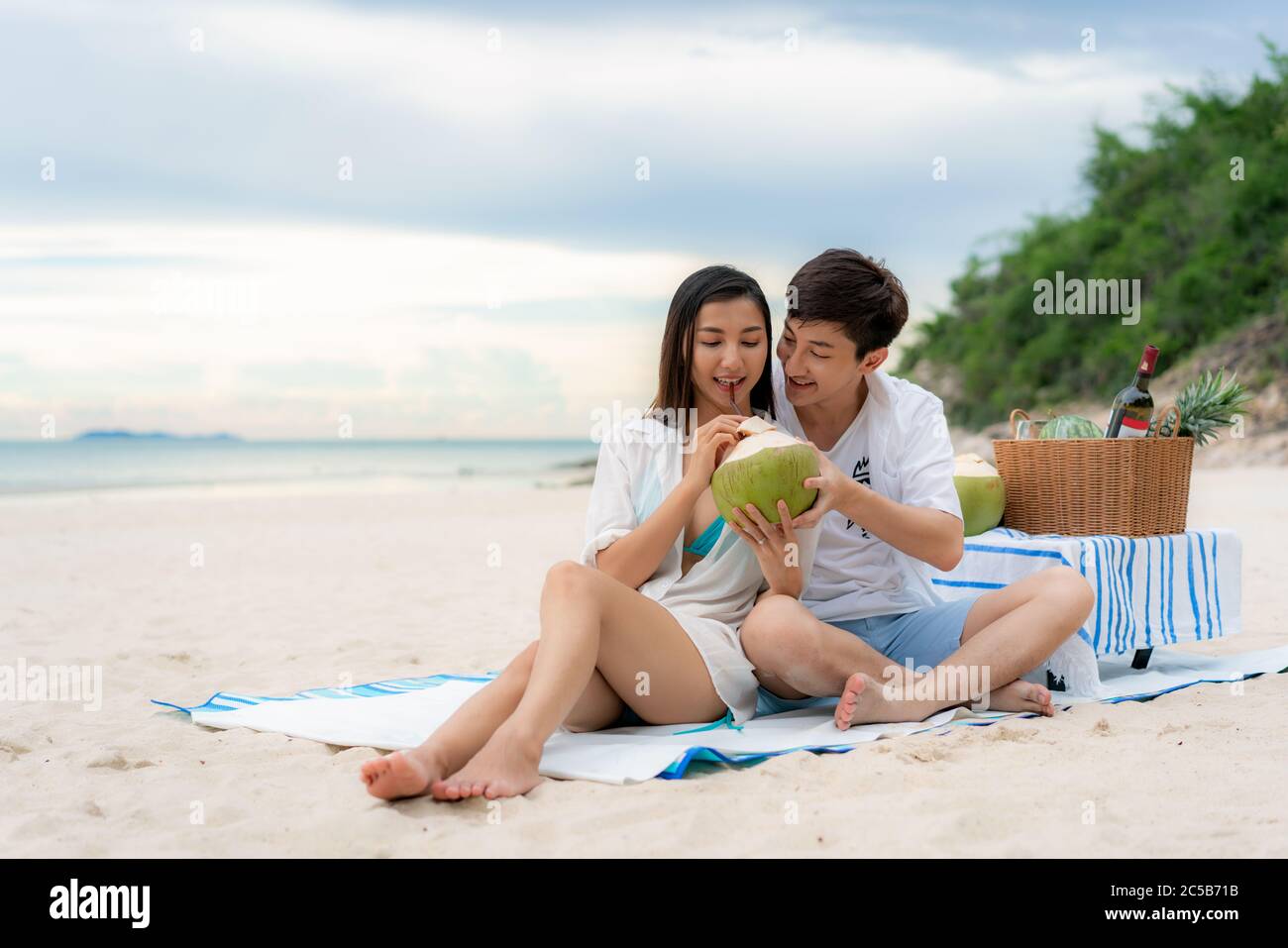 Asiatisches junges Paar auf der Picknickdecke sitzen und Kokoswasser am Strand und in der Nähe des Meeres mit tropischen Früchten im Hintergrund trinken. Sommer, holid Stockfoto