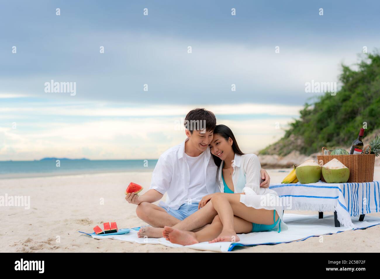 Asiatisches junges Paar, das auf der Picknickdecke sitzt und Wassermelone am Strand und in der Nähe des Meeres mit tropischen Früchten im Hintergrund isst. Sommer, Urlaub, Stockfoto