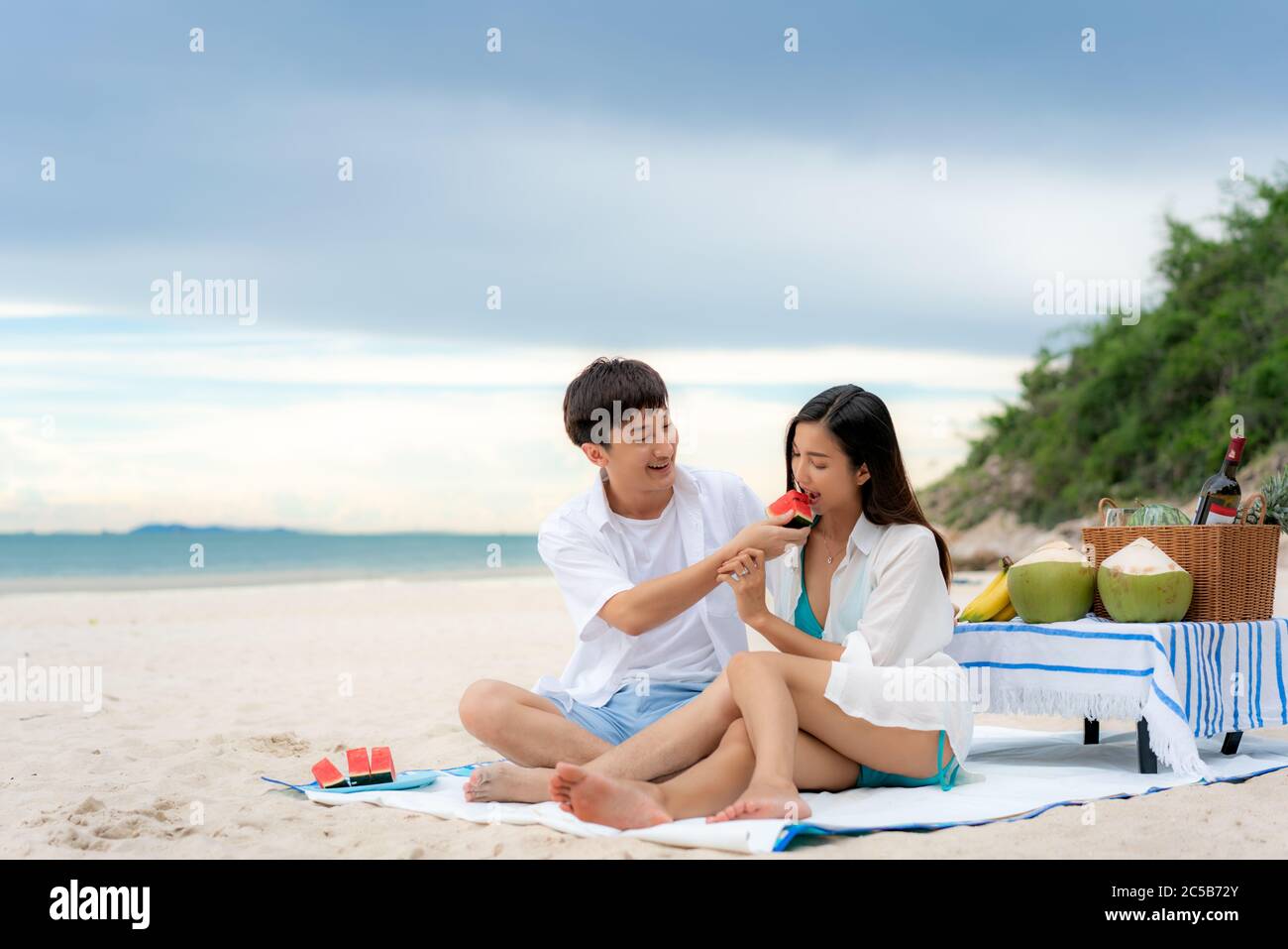 Asiatisches junges Paar, das auf der Picknickdecke sitzt und Wassermelone am Strand und in der Nähe des Meeres mit tropischen Früchten im Hintergrund isst. Sommer, Urlaub, Stockfoto