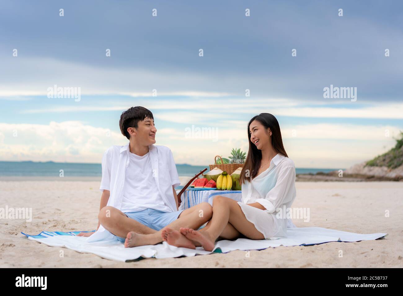 Asiatisches junges Paar sitzt auf der Picknickdecke und entspannen Sie sich am weißen Sandstrand und in der Nähe des Meeres mit tropischen Früchten im Hintergrund. Sommer, Urlaub, V Stockfoto