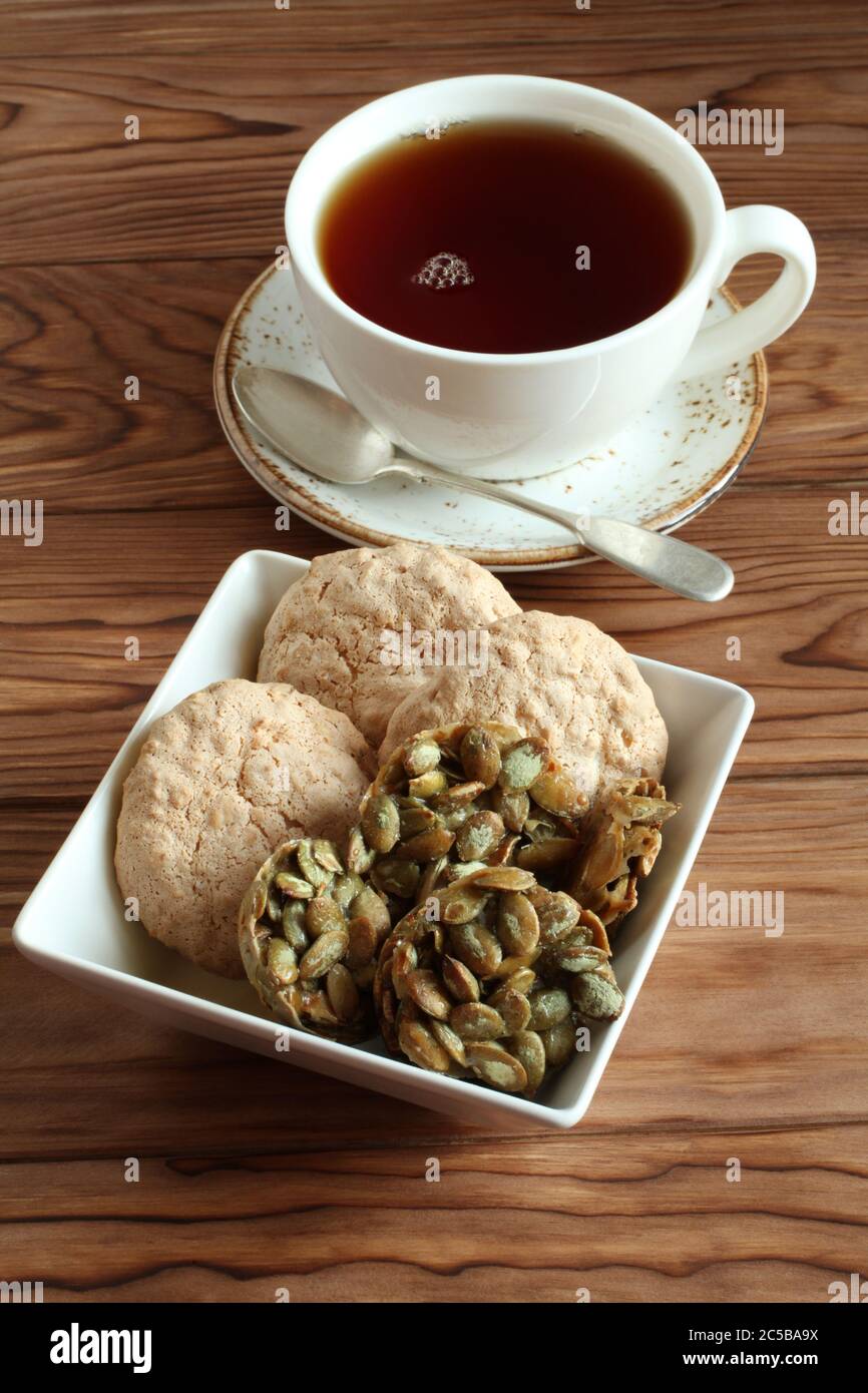 Mandelkekse und mit Zucker überzogene Kürbiskerne-Kekse in einer quadratischen Schüssel und eine Tasse schwarzen Tee auf einem Holztisch. Nahaufnahme Stockfoto