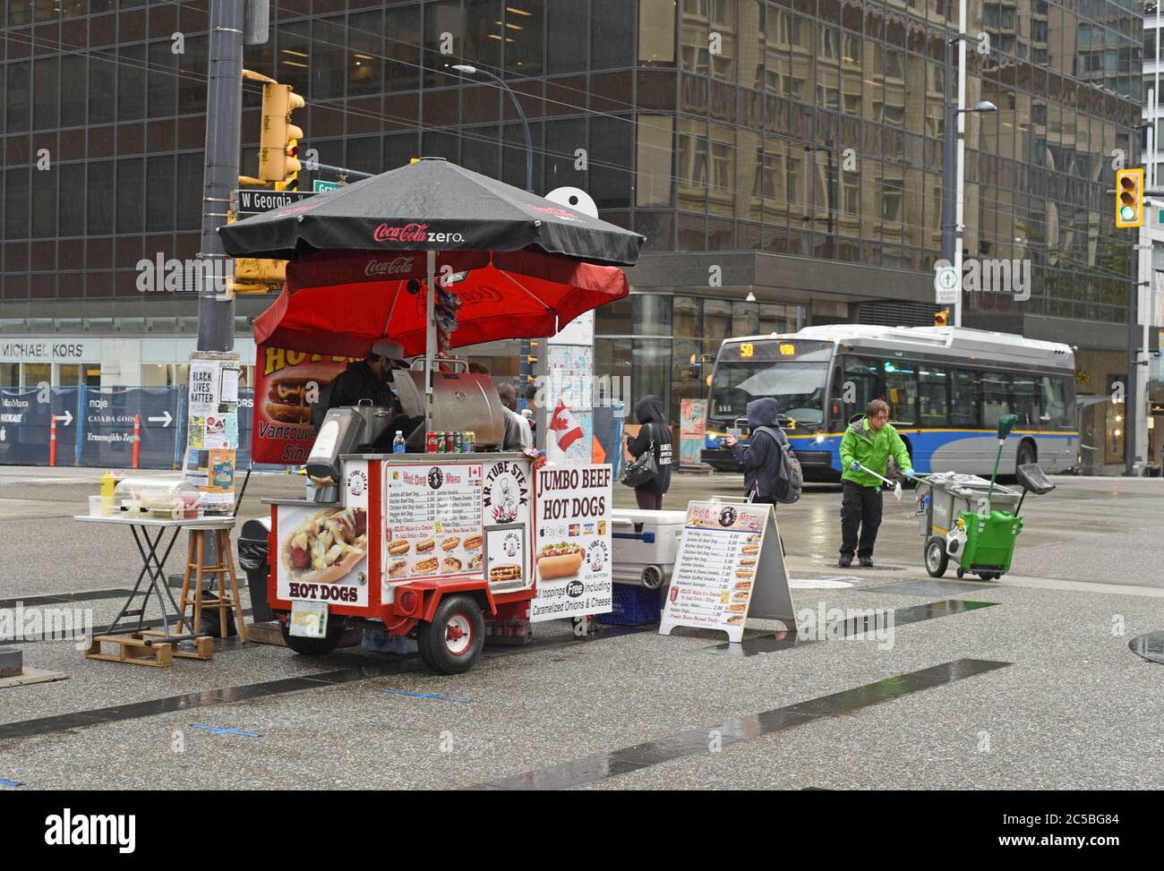 Ein Straßenreiniger arbeitet, als ein Bus hinter einem Hot Dog Food-Händler an der Ecke von Granville Street und W. Georgia Street in der Innenstadt von Vancouver, B Stockfoto