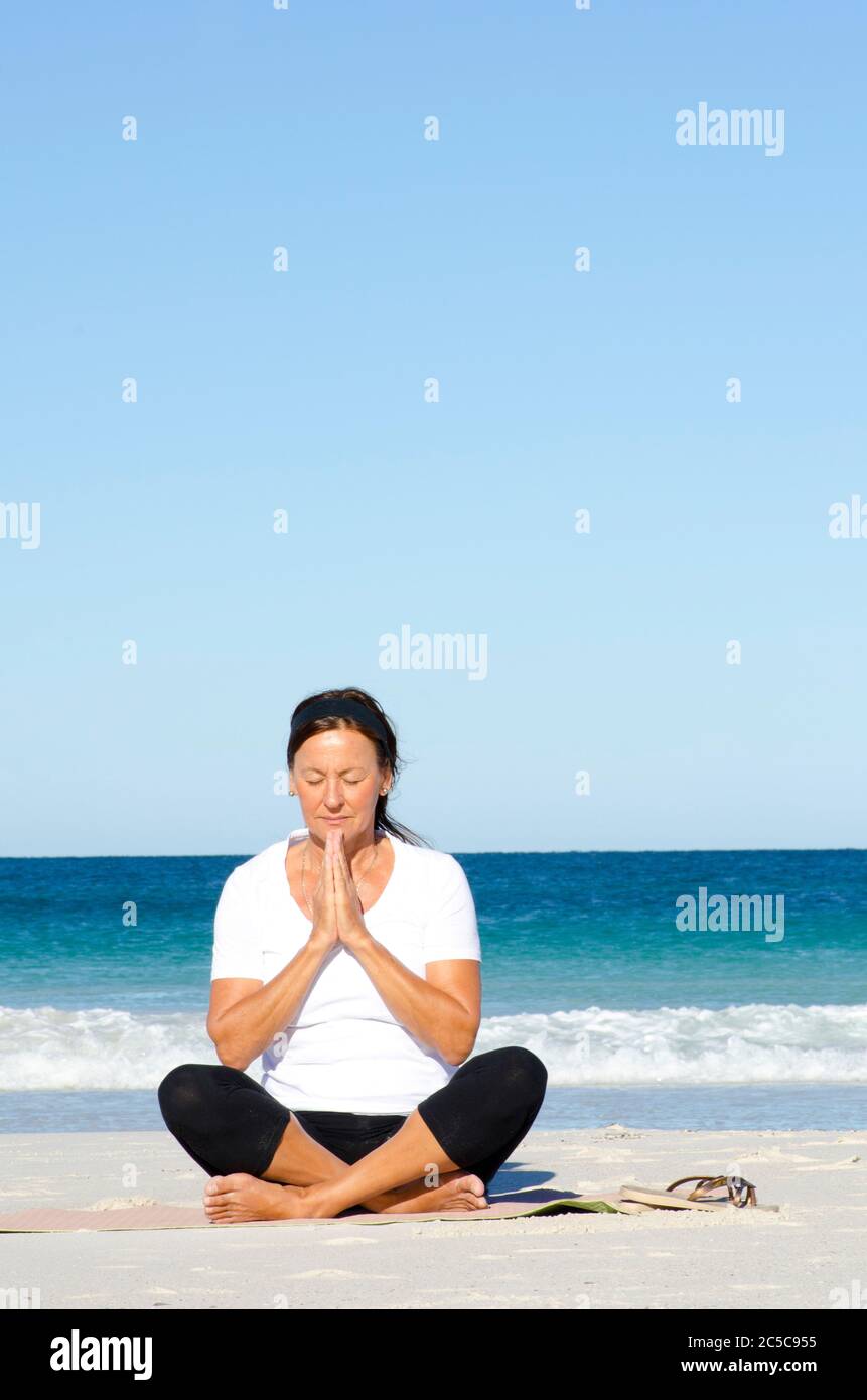 Attraktive und aktive ältere Frau, die sich mit Yoga und Meditation am Strand fit hält, isoliert mit Meer und blauem Himmel als Hintergrund und Kopierraum. Stockfoto
