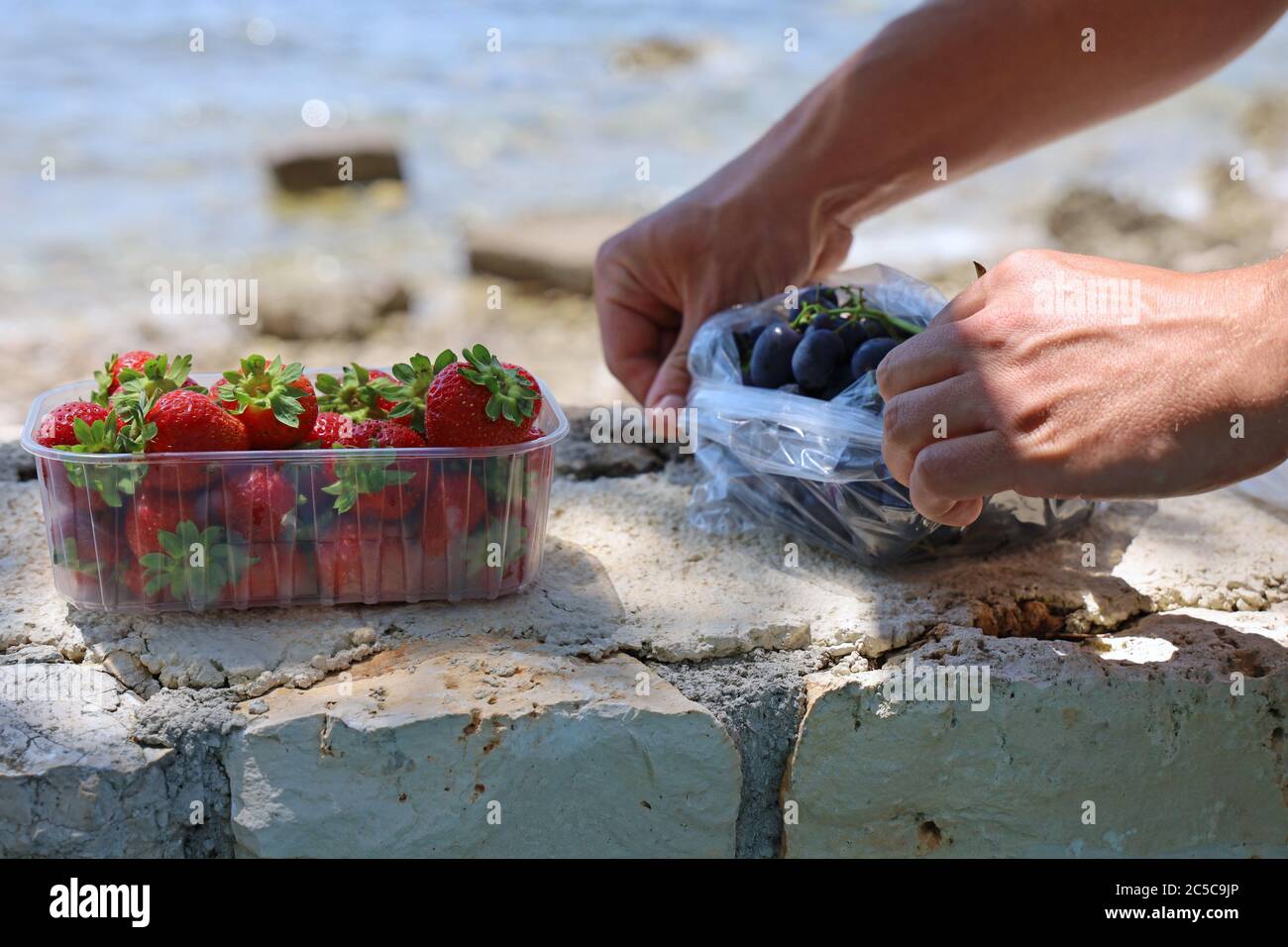 Saftige und reife Früchte an der Steinwand vor dem Meer, gesunde Ernährung am Strand, Nahaufnahme von männlichen Händen beim Auspacken von Trauben und Erdbeeren Stockfoto