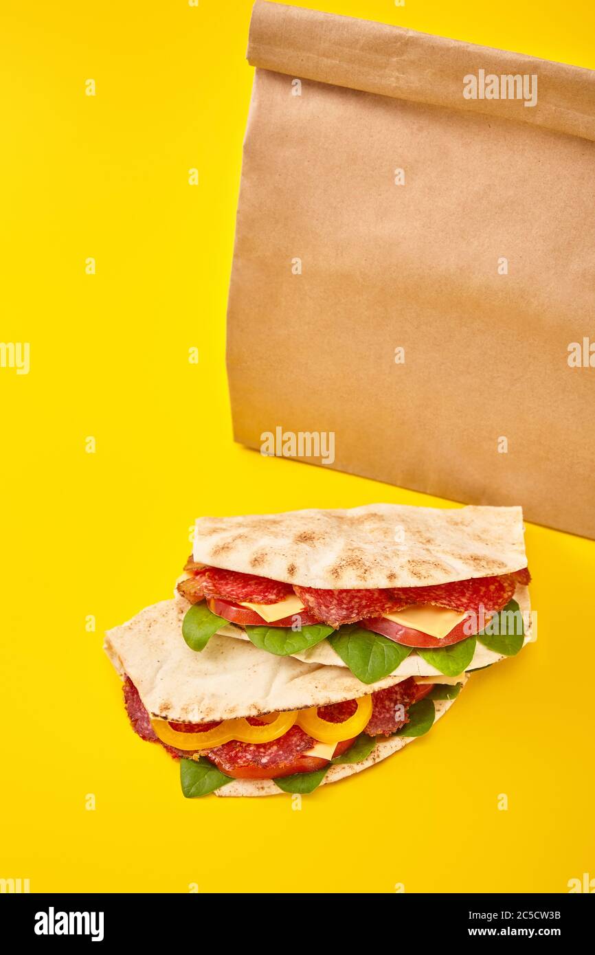 Frische Sandwiches mit Salami, Pita, Gemüse und Käse auf gelbem Hintergrund neben dem Takeaway-Beutel Stockfoto