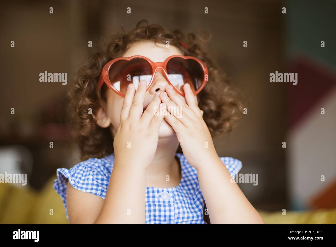 Portrait von hübschen Mädchen mit dunklen lockigen Haaren in Sonnenbrillen traumhaft den Mund mit den Händen zu Hause Stockfoto