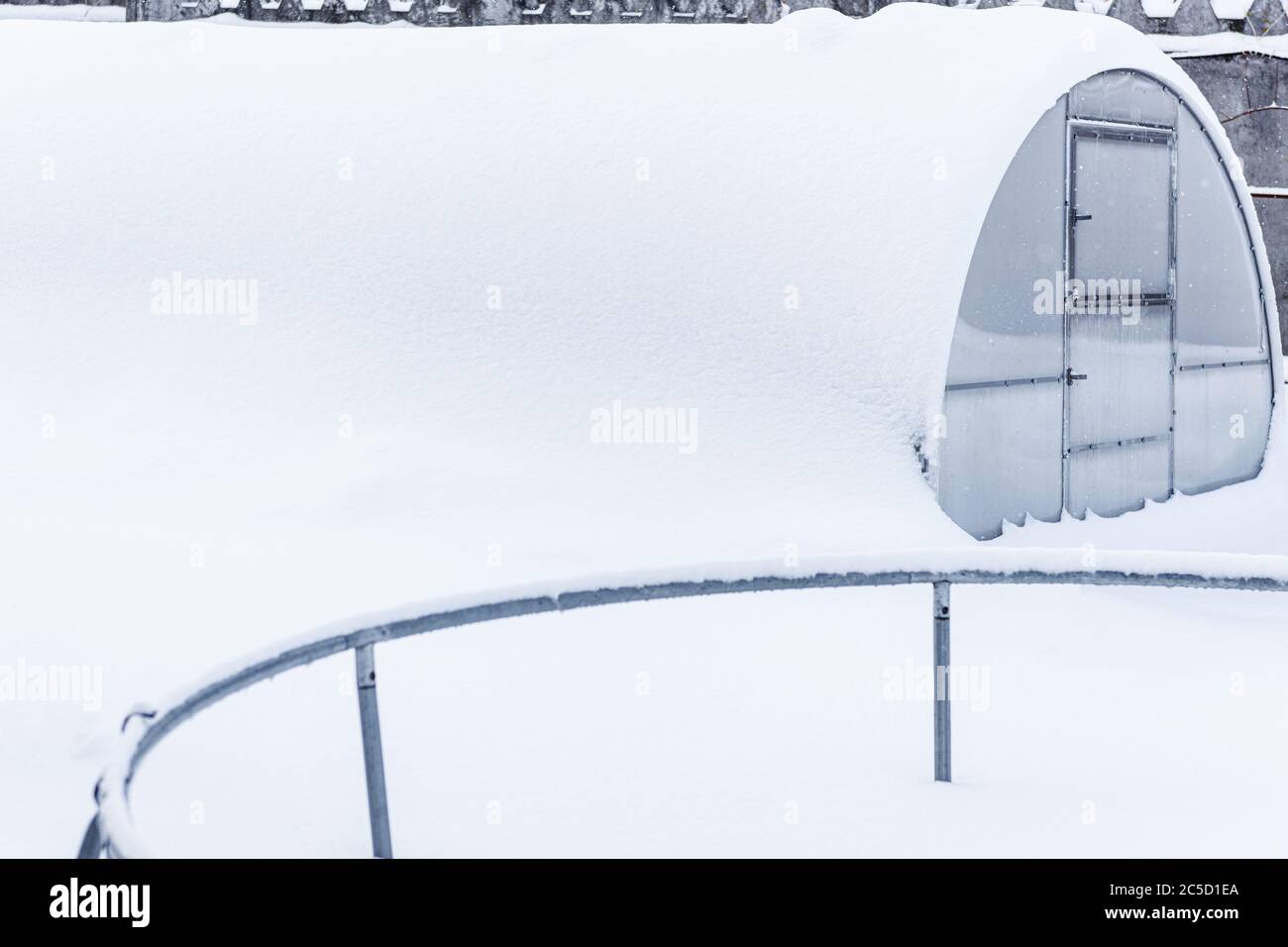 Winter, Schnee, Frost im Garten. Trampolin auf weißem Schnee in der Nähe des Gewächshauses. Gewächshaus im Schnee. Schneebedecktes Gewächshaus. Viel Schnee auf dem Dach Stockfoto