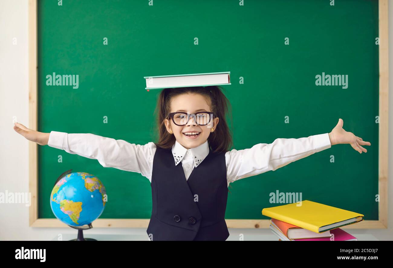 Zum ersten Mal zur Schule. Eine fröhliche Schülerin mit einem Buch über dem Kopf sitzt bei einer Lektion im Klassenzimmer. Stockfoto
