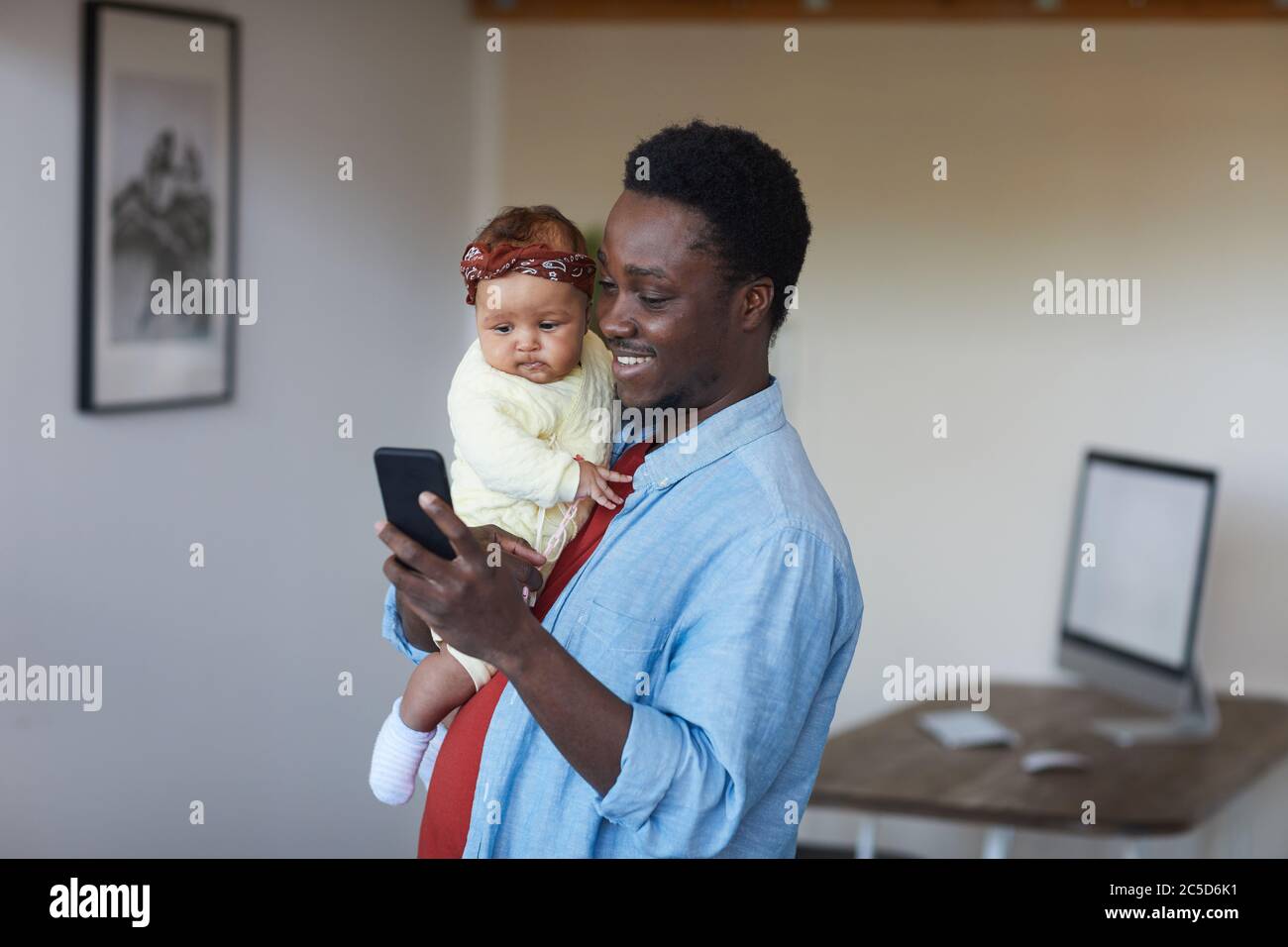 Afrikanisch lächelnder Vater hält ein Mädchen auf den Händen und benutzt sein Handy zu Hause Stockfoto