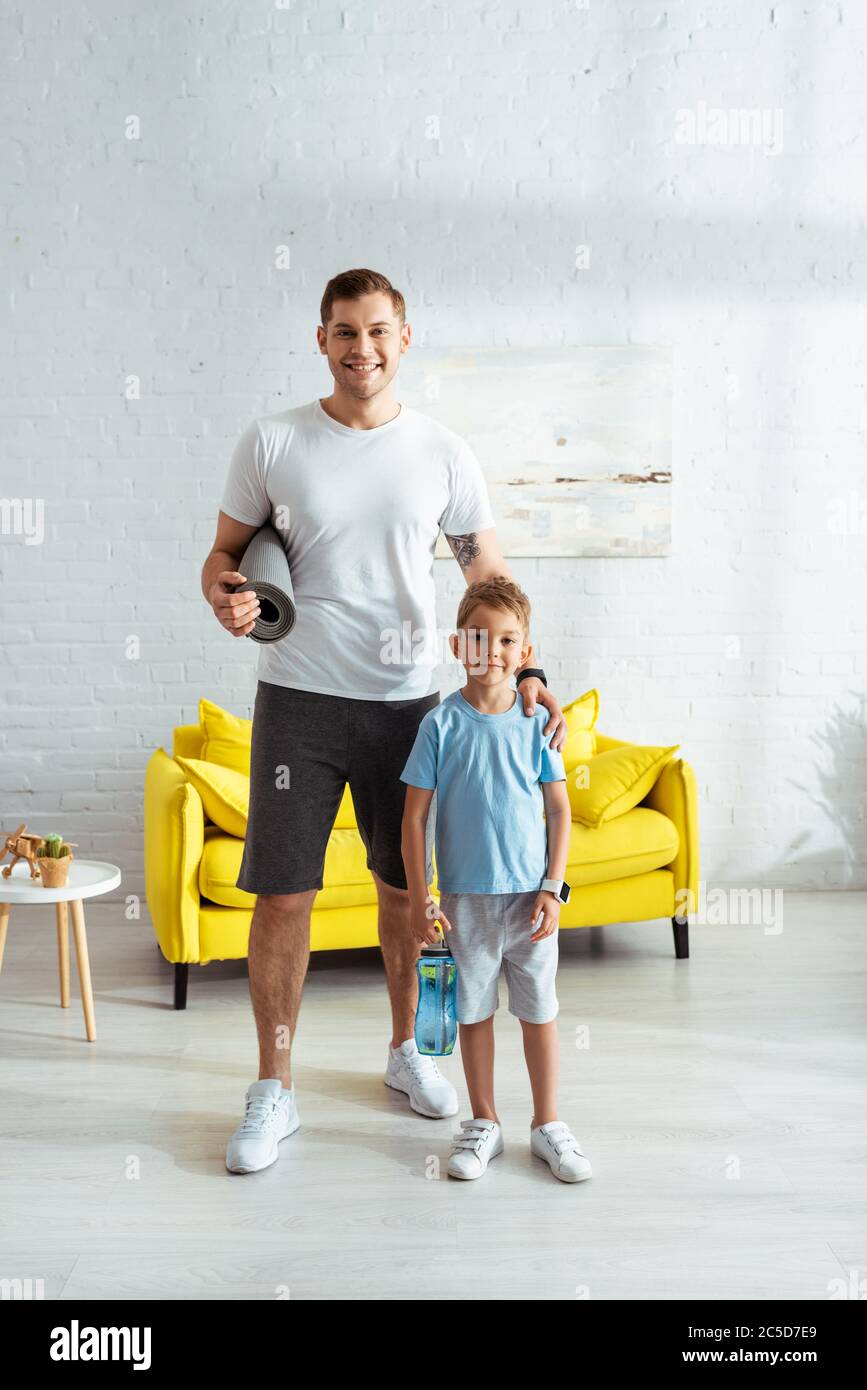 Lächelnder Mann mit Fitnessmatte, der die Schulter eines glücklichen Sohnes berührt, der eine Sportflasche hält Stockfoto