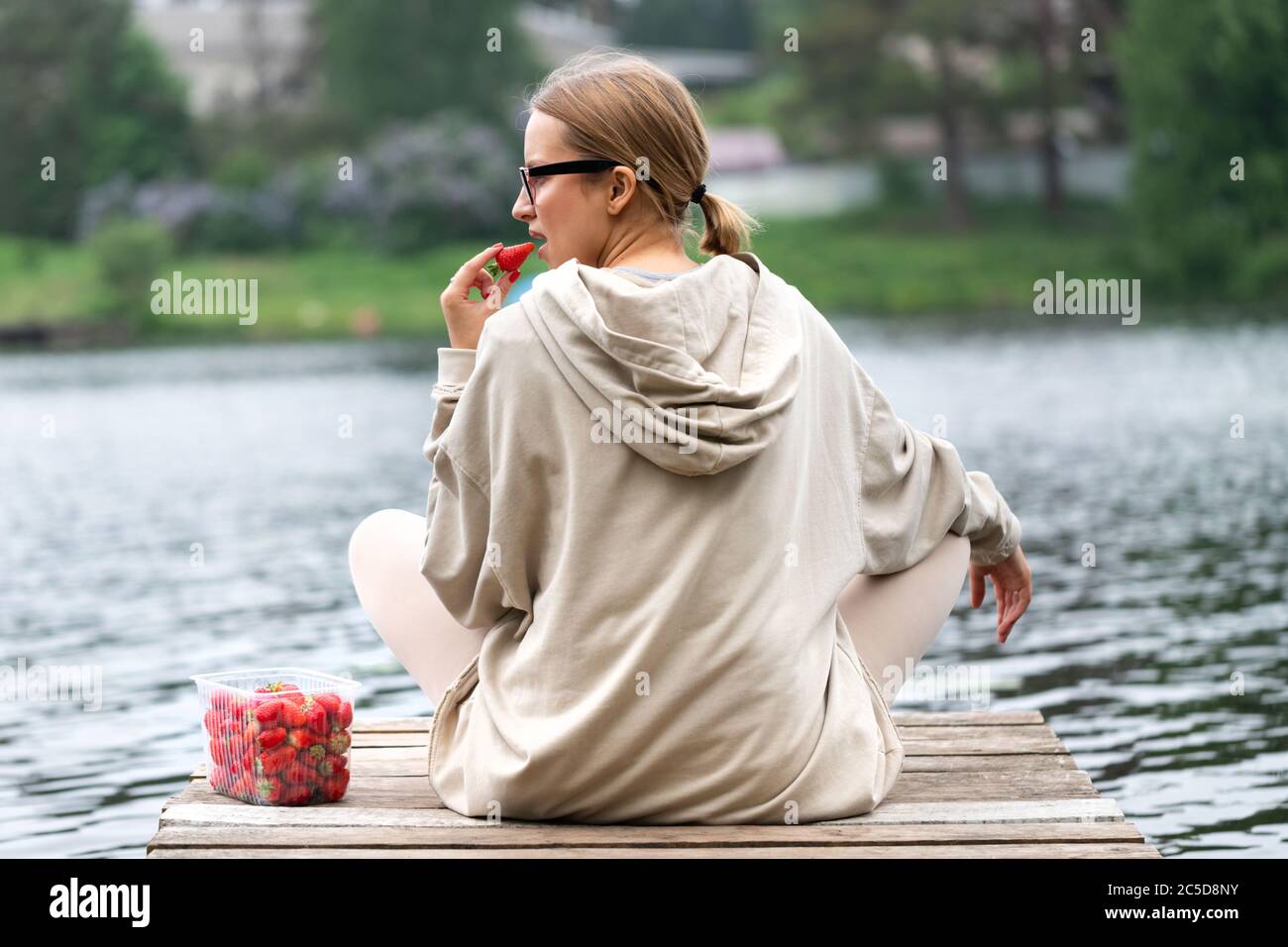 Junge Frau, die frische Beerenobst-Erdbeeren in Plastikbehälter isst, auf einem hölzernen Pier am Fluss ruht, Rückansicht. Sommerzeit. Die Freude des Landes lif Stockfoto