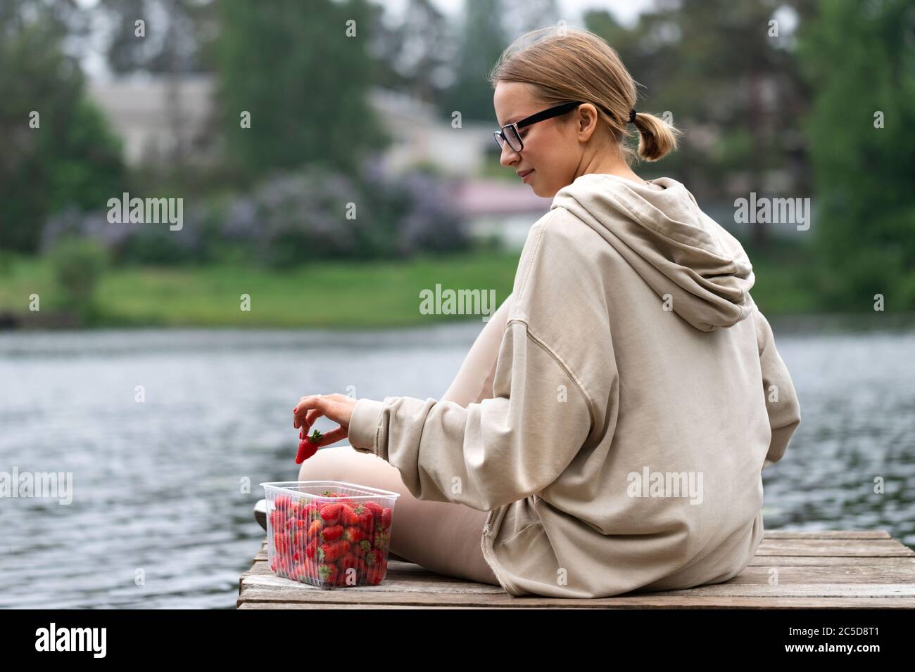 Junge Frau, die frische Beerenobst-Erdbeeren in Plastikbehälter isst, auf einem hölzernen Pier am Fluss ruht, Rückansicht. Sommerzeit. Die Freude des Landes lif Stockfoto
