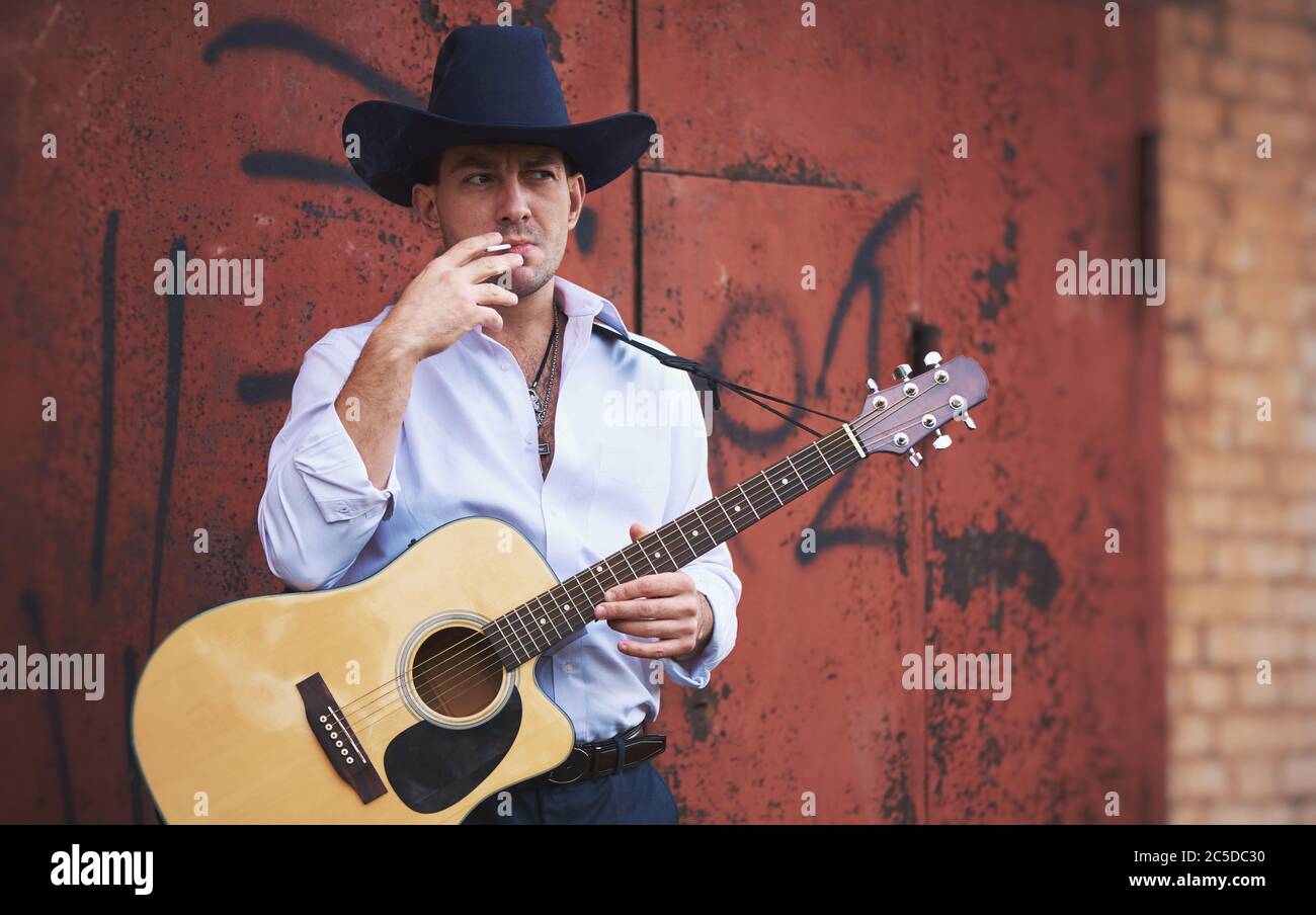 Der gutaussehende Mann trägt einen Cowboy-Hut, raucht eine Zigarette und hält eine akustische Gitarre Stockfoto