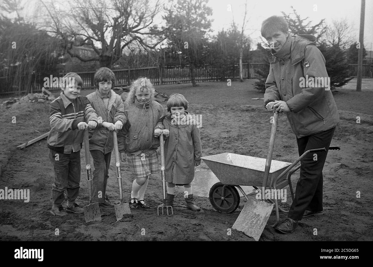 Neuer Schulgarten, vier Grundschüler mit ihrem männlichen Lehrer stehend, mit Pik, draußen in der Gegend, wo der neue Schulgarten sein wird, 1980er, England, Großbritannien. Outdoor-Aktivitäten wie Gartenarbeit und Pflanzen von Blumen und Gemüse haben sich als sehr erfolgreich erwiesen, um jungen Kindern zu helfen, sich sozial und geistig zu entwickeln und werden von Schulleiters aktiv gefördert, wenn Land und Raum zur Verfügung stehen. Stockfoto