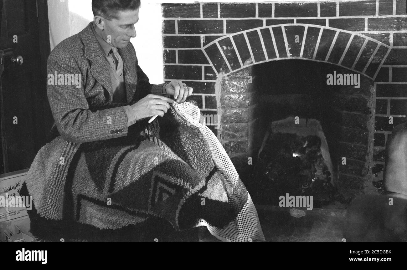 Mann, der eine Sportjacke mit Krawatte trägt, sitzt an einem offenen Kohlefeuer oder befestigt eine rutschfeste Gummiunterlage an einem gemusterten Wollteppich, England, Großbritannien, um 1940er Jahre. Die Unterlage würde auch die Lebensdauer des Teppichs erhöhen und helfen, kalte Böden zu isolieren und Ebene unebene. Stockfoto