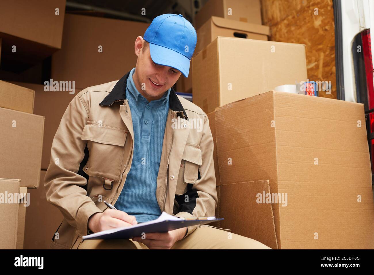 Der Zusteller füllt die Dokumente, während er draußen mit Kartons im Hintergrund sitzt Stockfoto