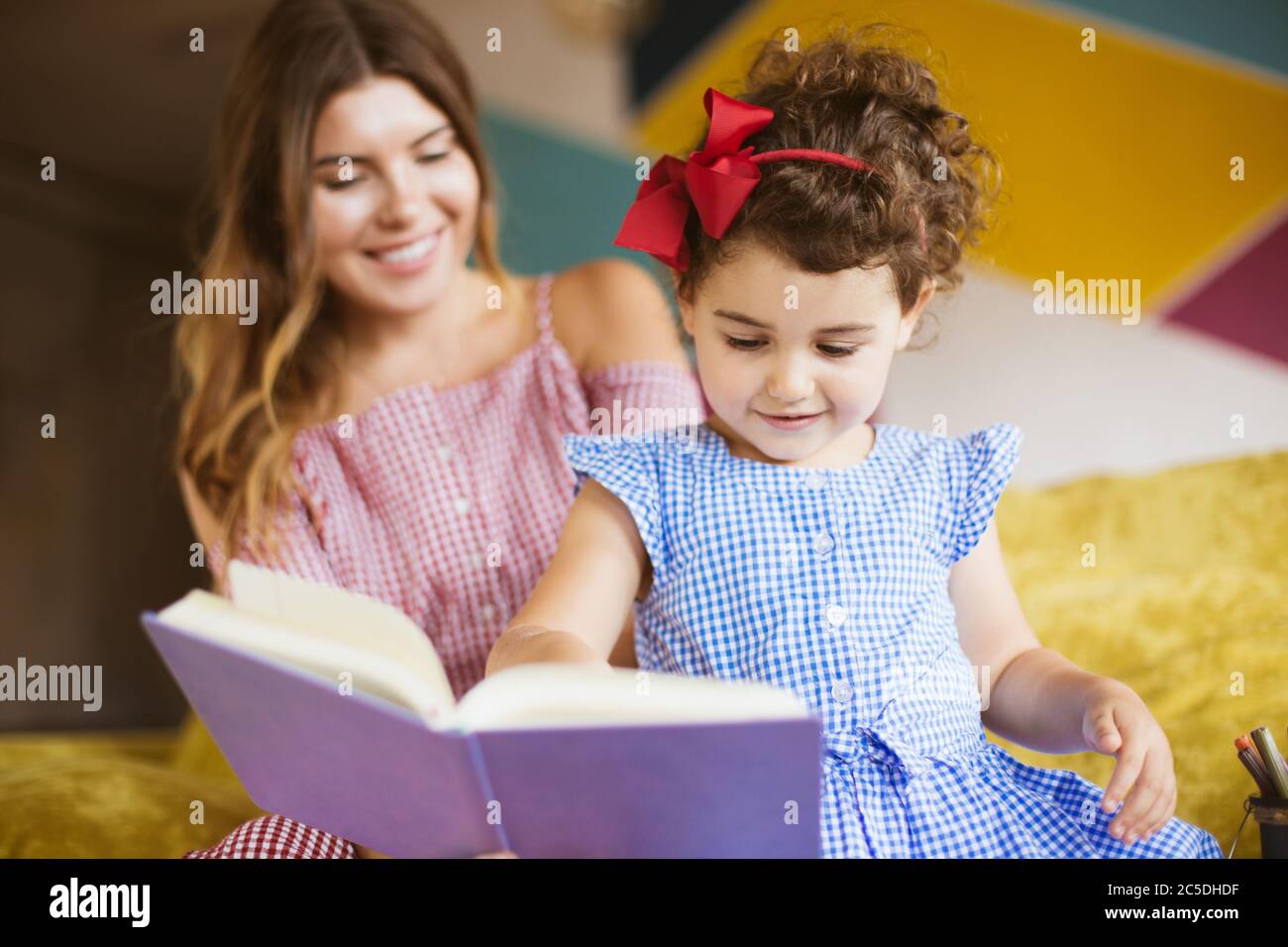 Hübsches kleines Mädchen mit dunklen lockigen Haaren im Kleid glücklich Kinderbuch mit Mutter auf Sofa zu Hause lesen Stockfoto