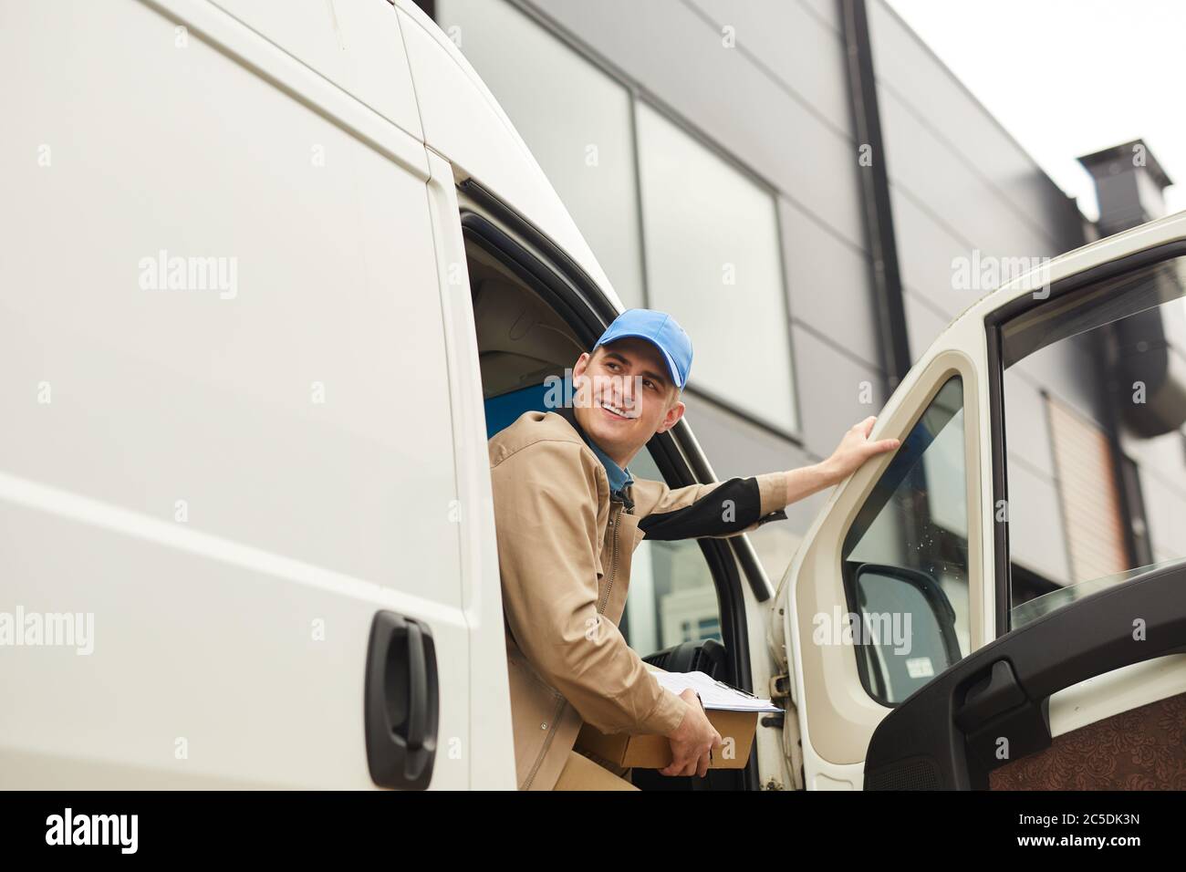 Junger lächelnder Kurier hält Paket, während er im Van sitzt, den er Pakete liefert Stockfoto