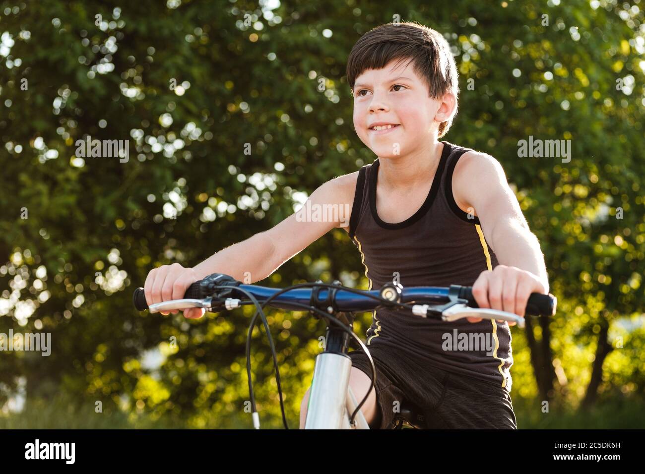 Glücklich gesunder Junge, der im Park in Nahaufnahme mit dem Fahrrad gegen grüne Bäume in einem aktiven Lifestyle-Konzept unterwegs ist Stockfoto