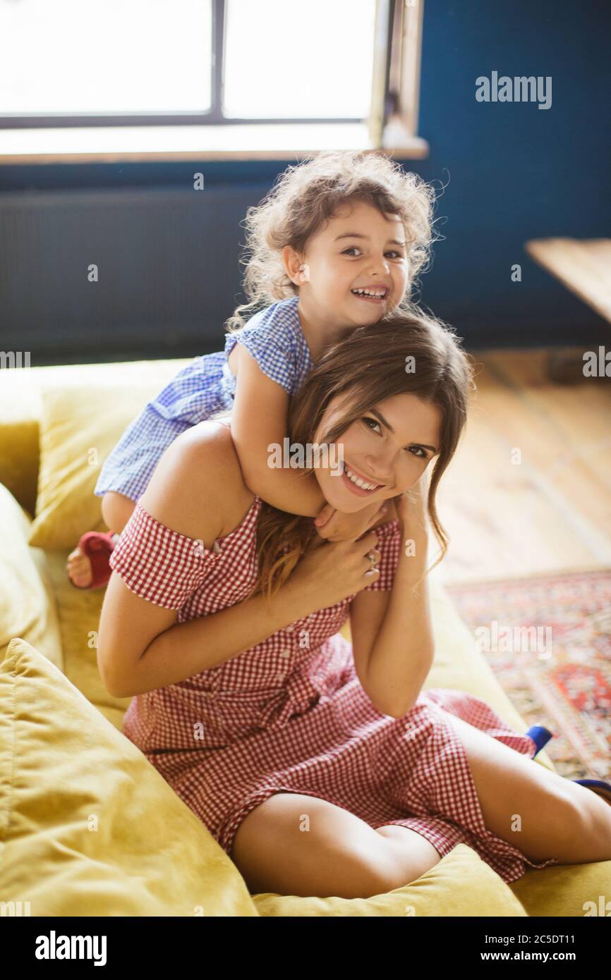 Fröhliche Baby-Mädchen mit dunklen lockigen Haaren umarmende Mutter glücklich in der Kamera suchen, während die Zeit zusammen auf dem Sofa zu Hause verbringen Stockfoto