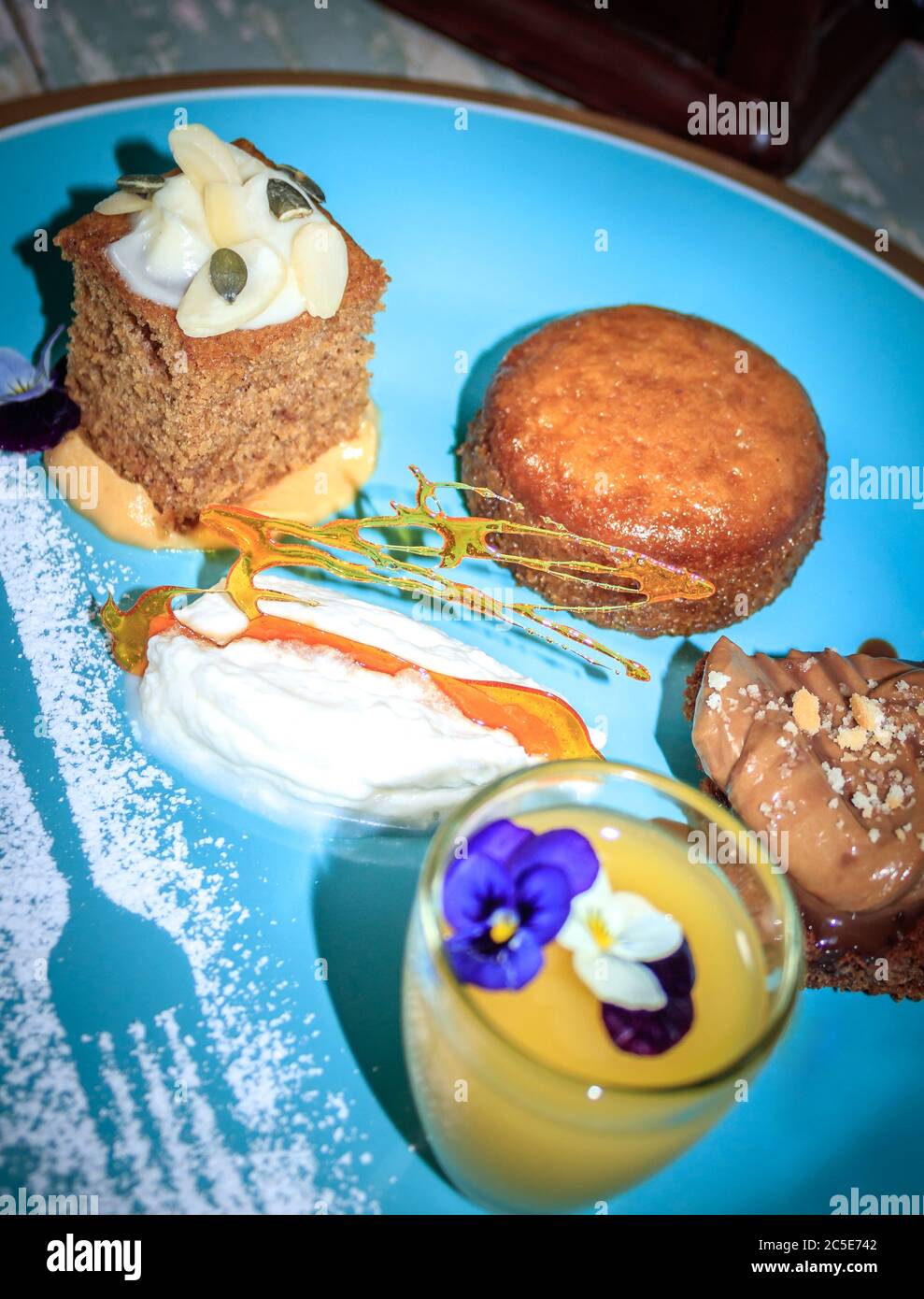 Schokoladenbrownie, malva Pudding, Kürbis-Gewürzkuchen mit glasierten Zuckerguss und Zitronenquark in einem kleinen Glas auf einem blauen Teller mit Puderzucker-Dekoration Stockfoto