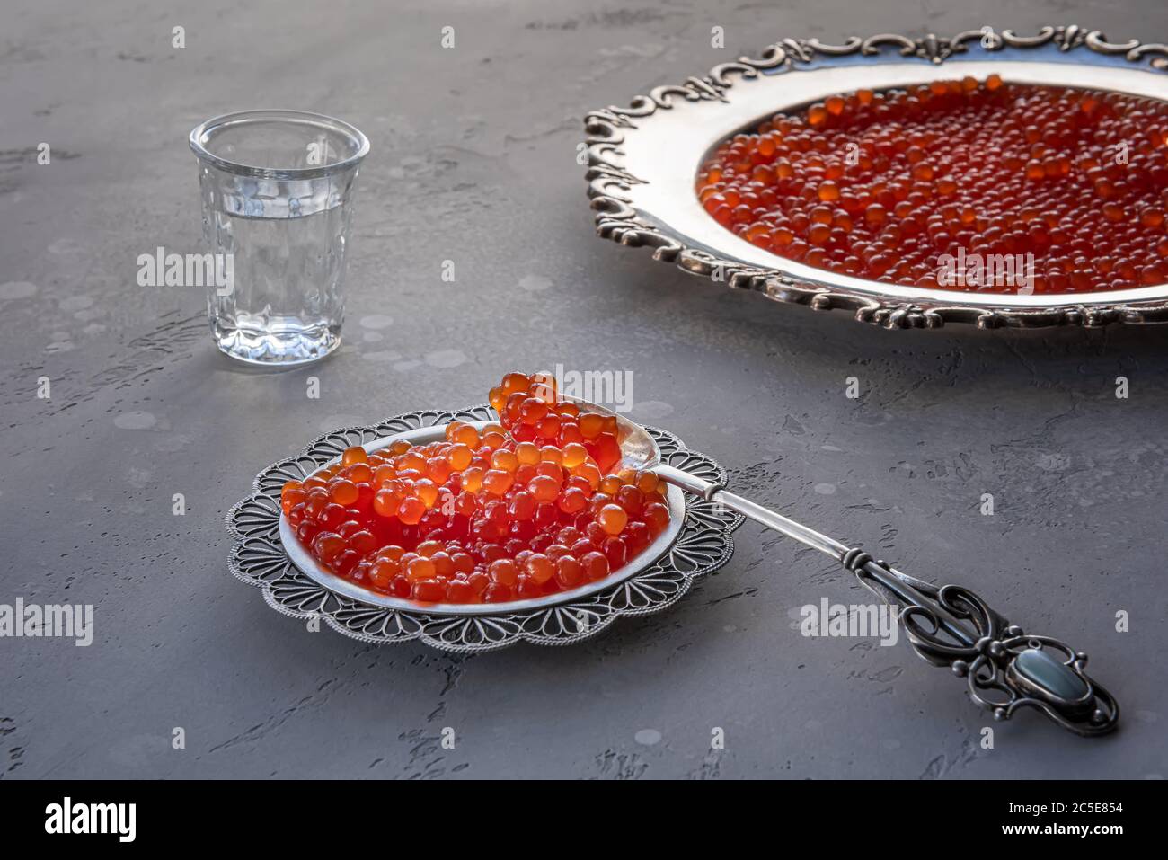Roter Kaviar in silbernem Löffel und Teller auf grauem Hintergrund. Wodka Schuss und Teller mit Kaviar auf unfokussierten Hintergrund. Selektiver Fokus. Gesunde Ernährung conce Stockfoto