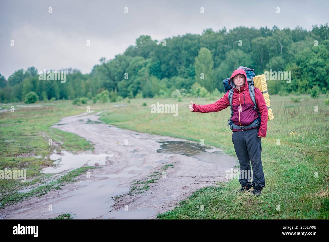 Guy Tourist Wanderer läuft entlang der Schotterstraße mit einem Rucksack in einer roten Jacke im Regen Stockfoto