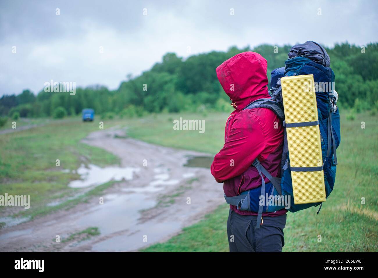 Guy Tourist Wanderer läuft entlang der Schotterstraße mit einem Rucksack in einer roten Jacke im Regen Stockfoto