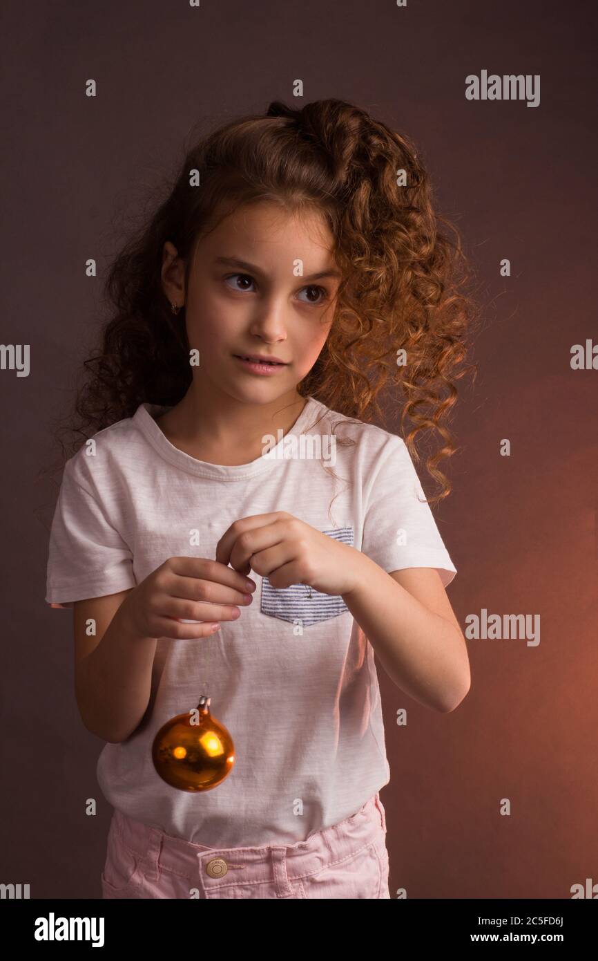 Ein kleines Mädchen in einem weißen T-Shirt mit lockigen langen Haaren, hält einen Ball von Weihnachtsbaum Dekoration, schaut weg auf einem dunklen Studio lila Hintergrund Stockfoto