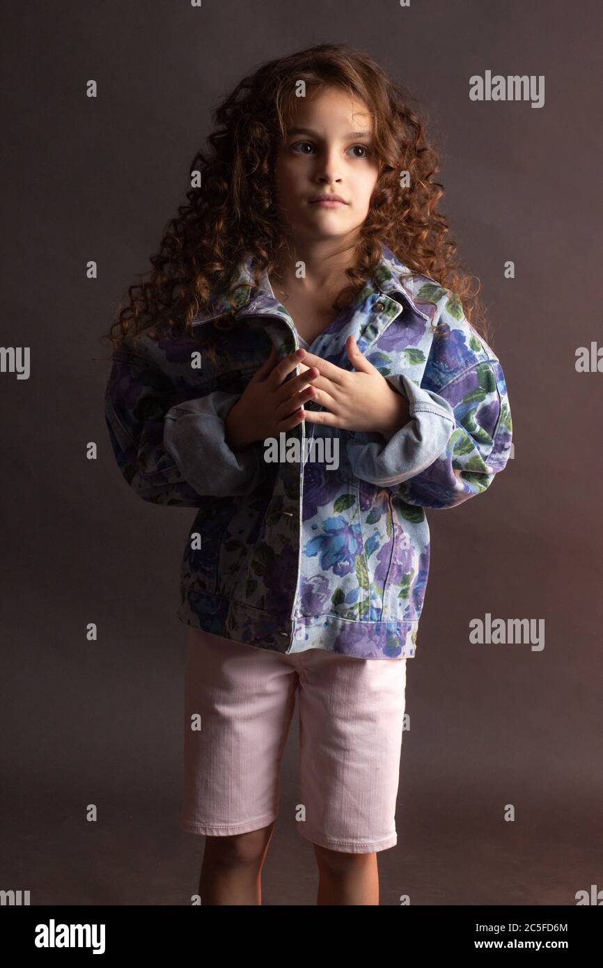 Studio-Portrait eines kleinen Mädchens mit langen lockigen Haaren, in einer bunten Jacke und leichten Shorts. Stockfoto