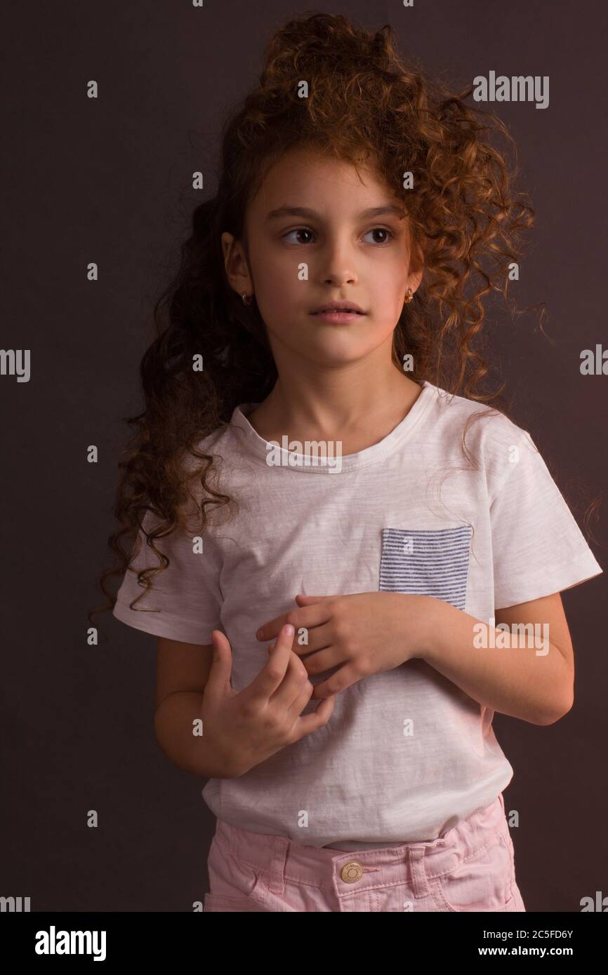 Ein kleines Mädchen in einem weißen T-Shirt mit lockigen langen Haaren, hält einen Ball von Weihnachtsbaum Dekoration, schaut weg auf einem dunklen Studio lila Hintergrund Stockfoto