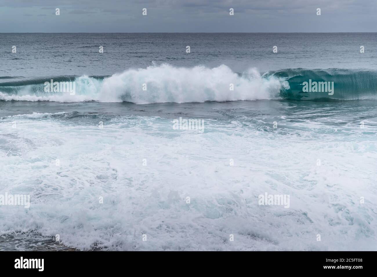 Wellen, die in ein felsiges Ufer voller großer Felsen stürzen und das Wasser brechen, das mit einem Paar Boote am Horizont auf See zu den Seiten springt Stockfoto