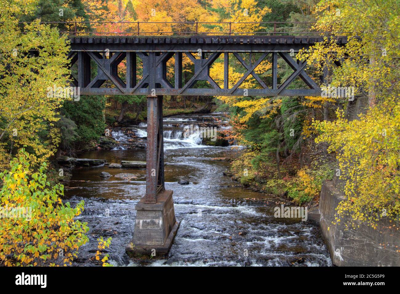 Eisenbahnbrücke über einen Fluss mit einem Wasserfall im Hintergrund, umgeben von lebhaften Herbstfarben in einer kleinen Stadt auf der Upper Peninsula in Michigan. Stockfoto