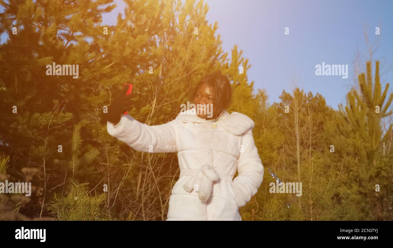 Schöne schwarze Frau in Pelzmantel macht Selfie mit Smartphone gegen wilden Winter immergrünen Wald unter klarem blauen Himmel Stockfoto