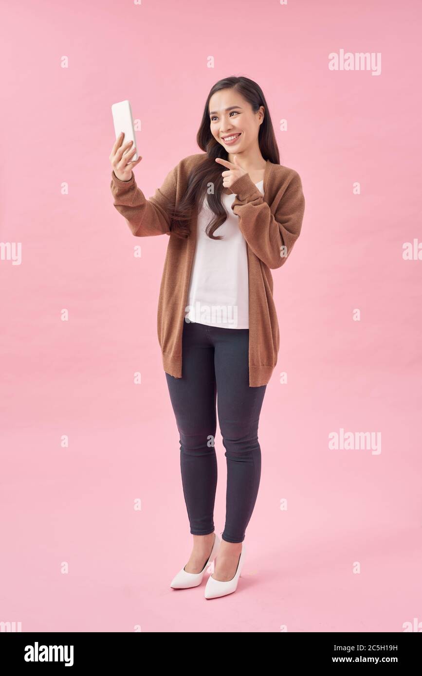 Glückliche junge asiatische Frau lässig gekleidet stehend isoliert über rosa Hintergrund, mit Handy Stockfoto