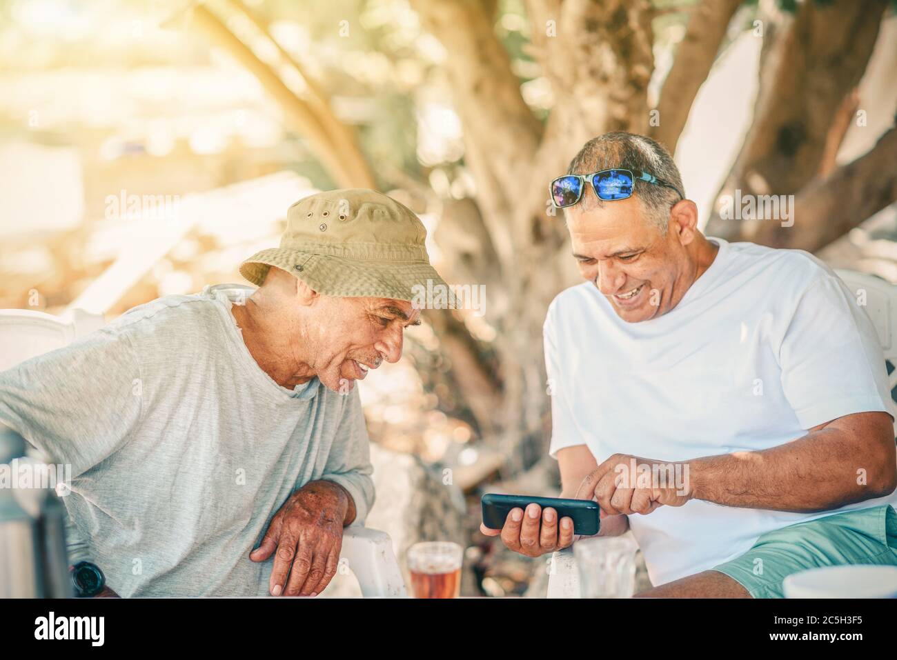 Zwei positive lächelnde Silber Surfer Senior Mann verwenden Smartphone und Tee trinken. Aktive gesunde Senioren Boomer Spaß im Garten. Stockfoto