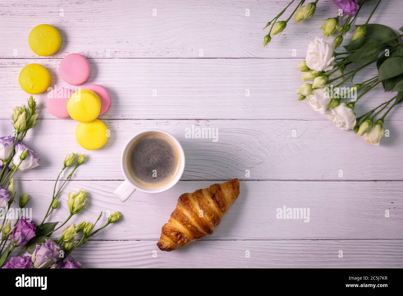 Makronen und Croissant mit Blumen und Tasse Kaffee auf weißem Holzhintergrund mit Kopierfläche. Draufsicht Stockfoto