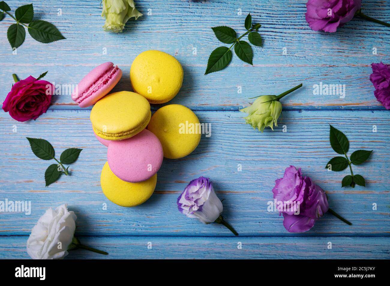 Rosa und gelbe Macarons mit Blumen auf blauem Holztisch. Draufsicht Stockfoto