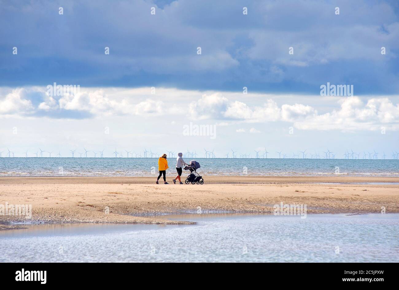 Pärchen schieben Kinderwagen am Strand entlang an einem kalten Tag im Sommer mit Windpark im Hintergrund Stockfoto