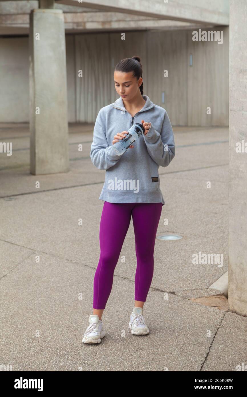 Eine Frau, die in violetten Leggings, grauem Sweatshirt und mit einer Getränkeflasche steht Stockfoto