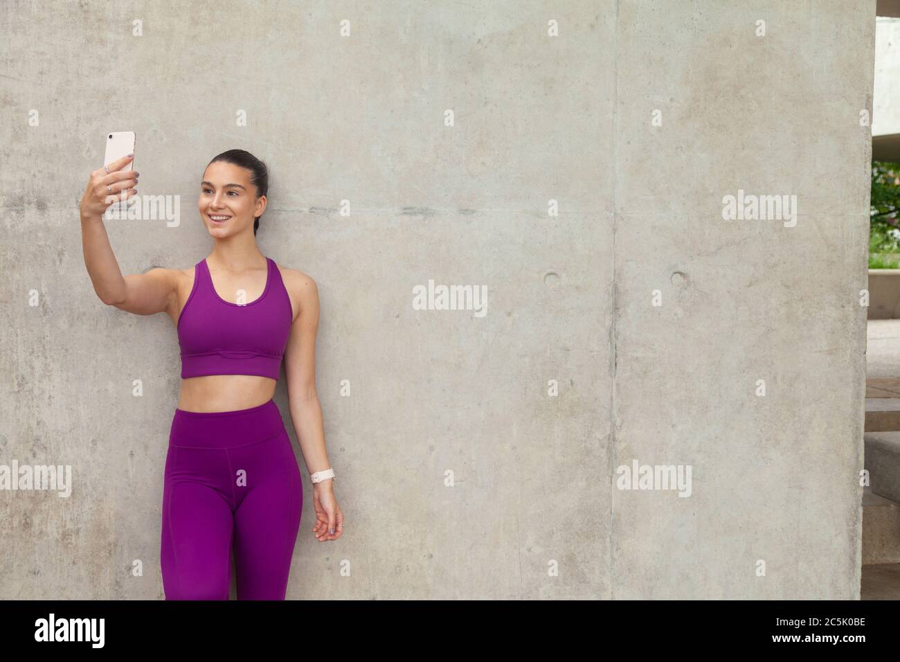 Eine attraktive Frau, die Sportkleidung trägt, die sich an eine Betonwand lehnt und ein Mobiltelefon hochhält. Stockfoto
