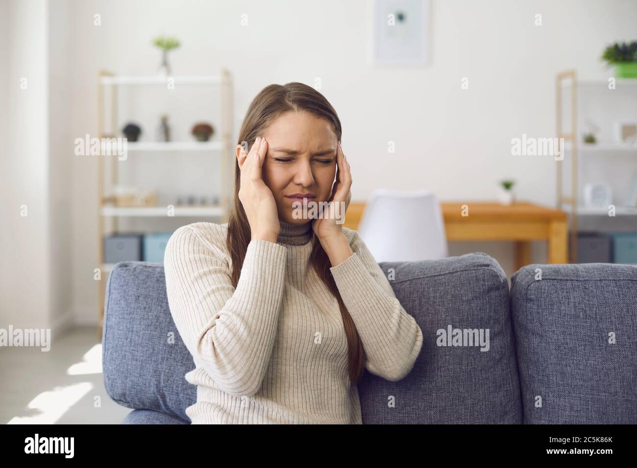 Kopfschmerzen. Migräne. Eine blonde Frau hat Kopfschmerzen, während sie auf einem Sofa in einem Raum sitzt. Stockfoto