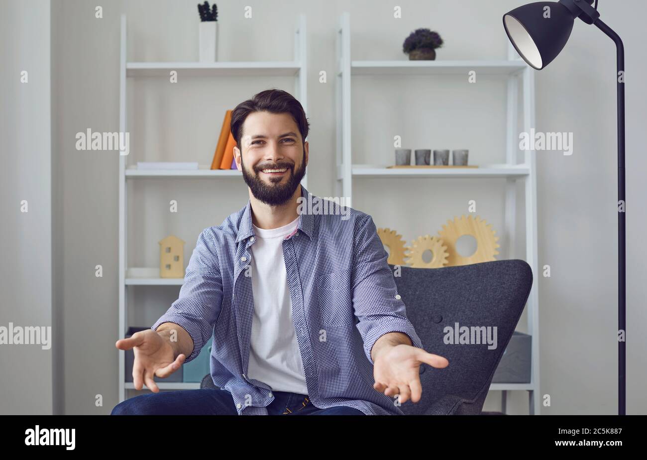 Junge kaukasische Mann Blick auf Kamera Video Chat mit offenen Armen in freundlichen Willkommensgruß Geste im Wohnzimmer. Stockfoto