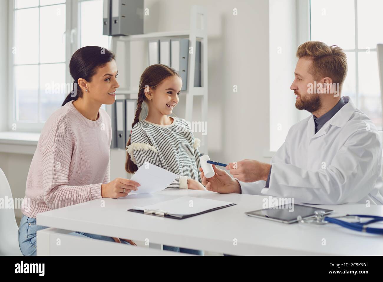 Der männliche Arzt gibt Mutter und Tochter ein Rezept, während er an einem Tisch in einem Klinikbüro sitzt. Stockfoto